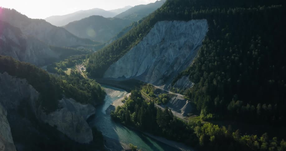 A cinematic drone shot flies past trees to reveal a breathtaking view of a deep mountain gorge with a turquoise river flowing through it at sunrise
