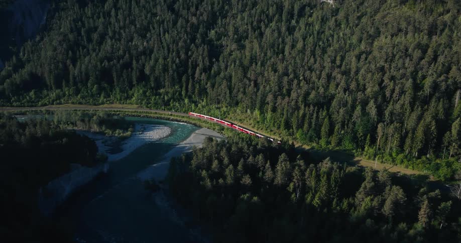 An aerial view shows a red passenger train traveling alongside a winding river through a dense, sunlit evergreen forest