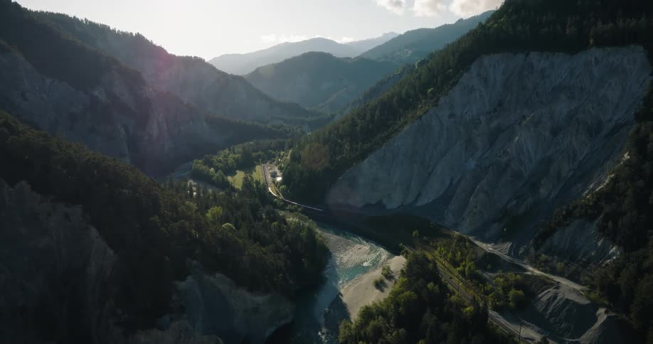 A scenic aerial view shows a red train traveling alongside a turquoise river through a stunning mountain valley in the Swiss Alps