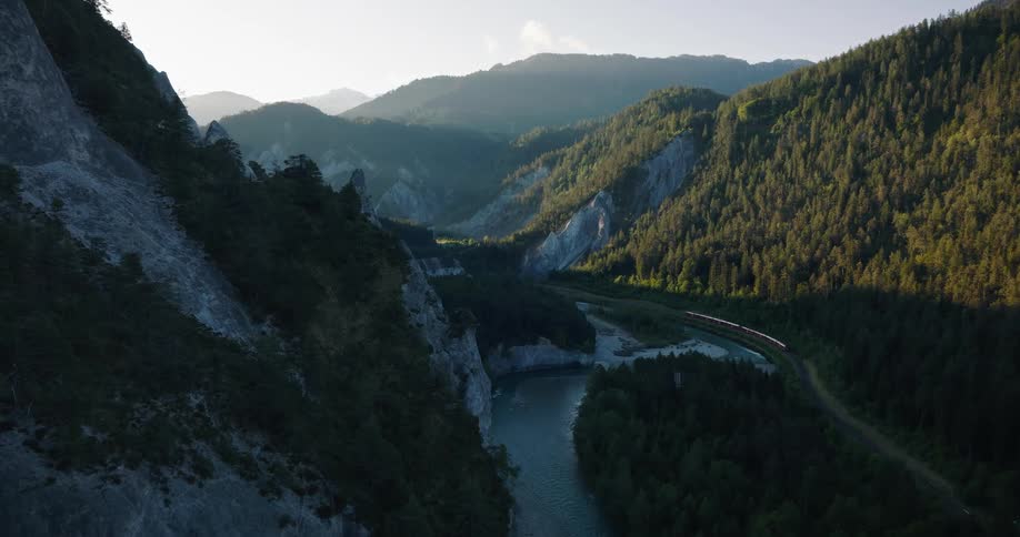 Aerial view of a red train traveling alongside a river through a majestic mountain valley during a golden sunrise