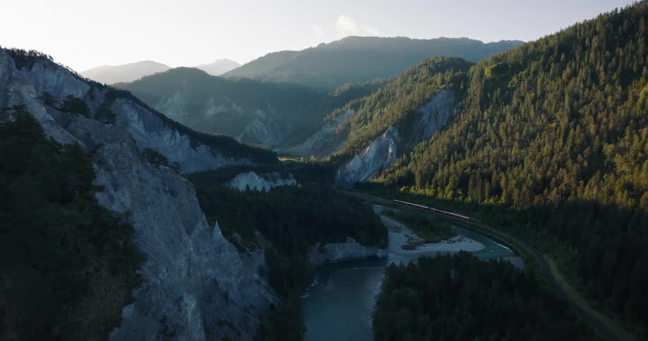 An aerial view of a red train traveling through a stunning mountain valley alongside a winding river at sunrise