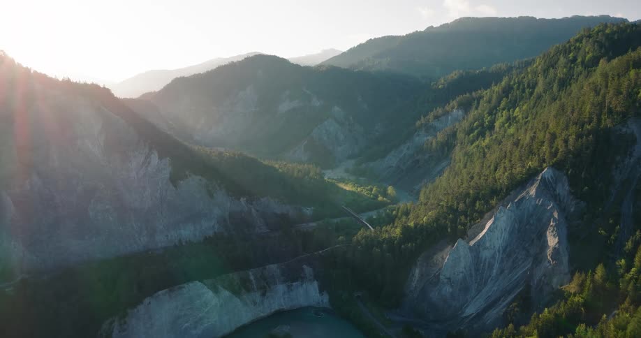 A stunning aerial view shows a train winding through a lush green and rocky mountain valley alongside a river during a beautiful sunrise