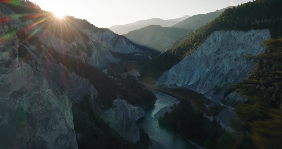 An aerial shot pushes forward past evergreen trees to reveal a stunning mountain gorge with a river below as the sun rises over the peaks