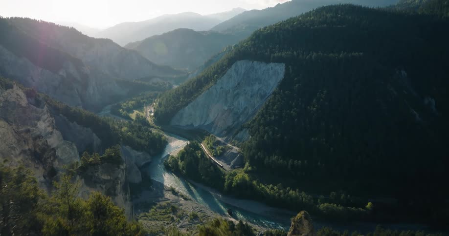 A majestic, wide shot captures a train journeying through the scenic Rhine Gorge in Switzerland, nestled between forested mountains and a winding river