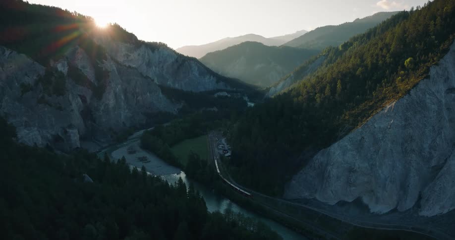 The sun sets over a beautiful mountain gorge as a red train travels alongside a winding river in a stunning aerial view