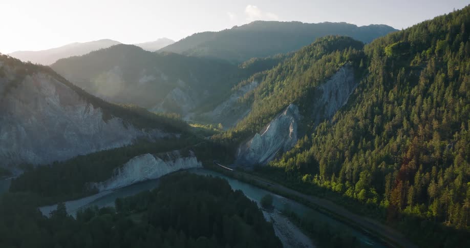 A red passenger train travels through a majestic mountain valley alongside a winding river during a golden sunrise