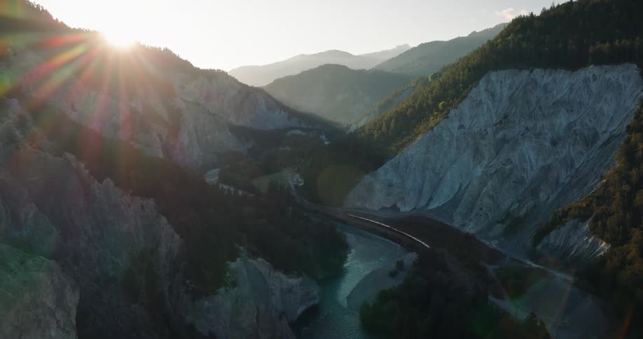 A scenic train travels along a river through a majestic mountain canyon at sunrise