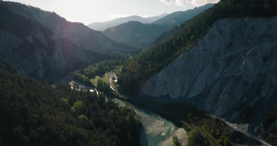 A majestic, static aerial view of the Rhine Gorge in Switzerland, where a turquoise river carves its way through a deep, forested valley at sunrise
