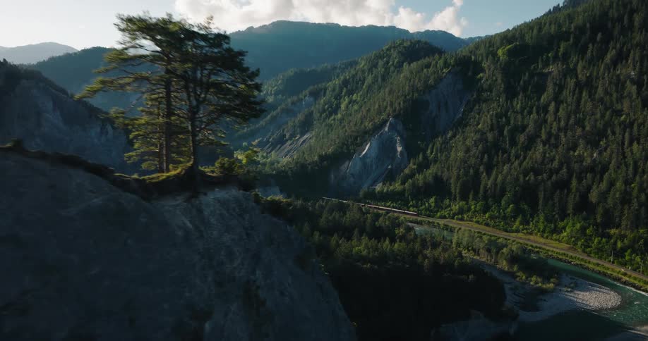 Epic aerial footage reveals a scenic train journeying through a breathtaking mountain valley alongside a winding river at golden hour
