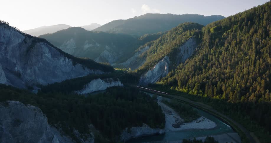 An aerial view shows a red train traveling alongside a river through a magnificent mountain valley at sunrise