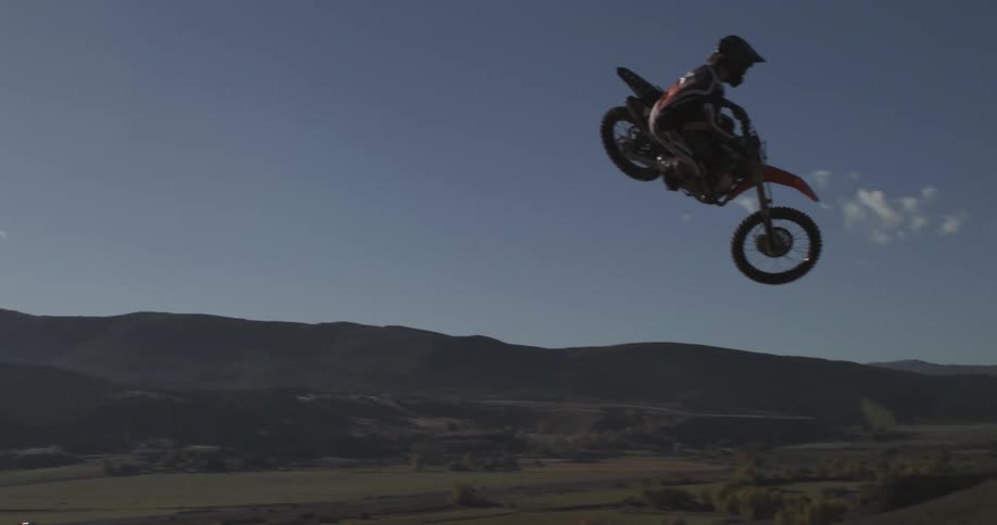 A motocross rider flies through the air in super slow motion against a backdrop of rolling hills and a clear blue sky