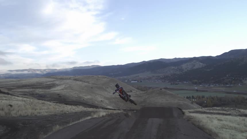 A motocross rider on a dirt bike soars through the air in super slow-motion after hitting a large dirt jump in a scenic, hilly landscape