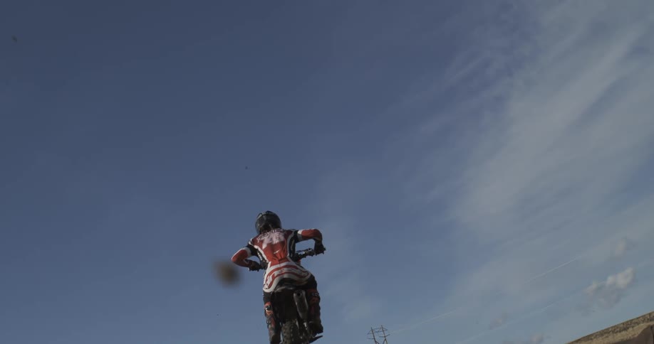 A motocross rider soars through the air in extreme slow motion, captured from a dramatic low angle against the blue sky