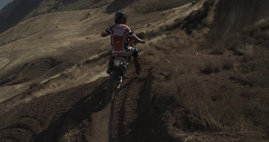 A motocross rider in full gear begins to ride down a steep dirt track on a hillside in slow motion