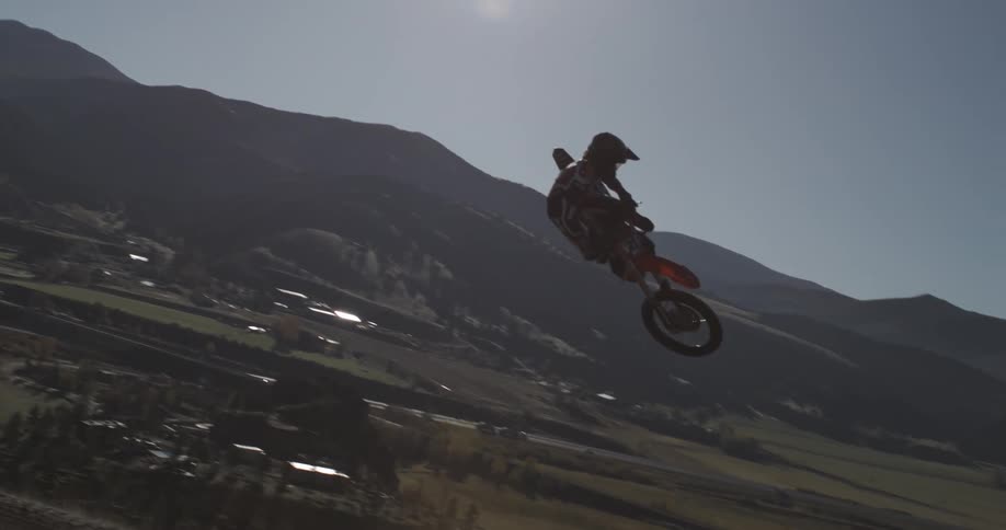 A silhouetted motocross rider soars through the air in dramatic slow motion with a scenic mountain valley below
