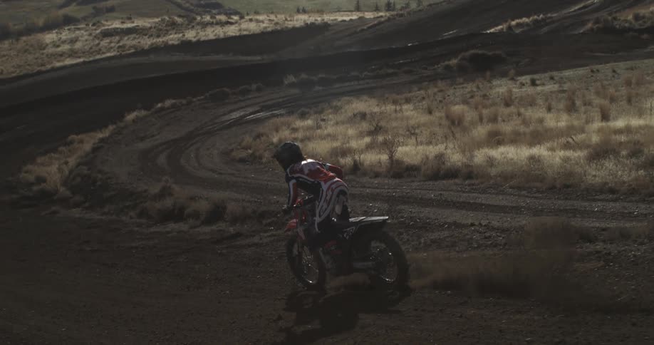 A motocross rider gracefully navigates a turn on a dirt track in slow motion, backlit by the brilliant sun over a vast rural landscape