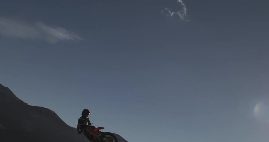 A motocross rider soars through the air in epic slow motion with a mountain backdrop and a bright sun flare