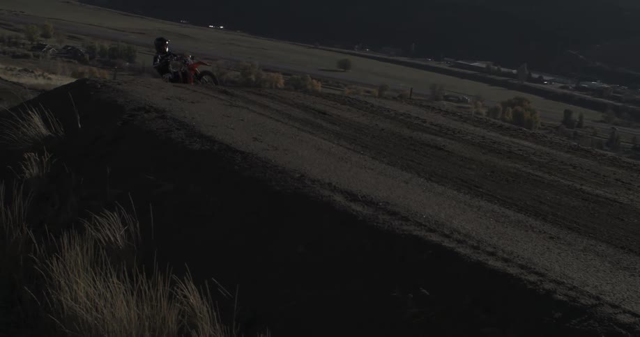 A motocross rider performs a slow-motion wheelie on a dirt track overlooking a valley at sunset