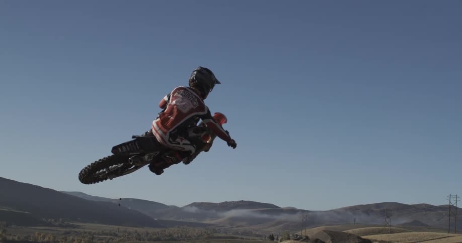 A motocross rider launches off a dirt ramp in super slow motion against a clear blue sky