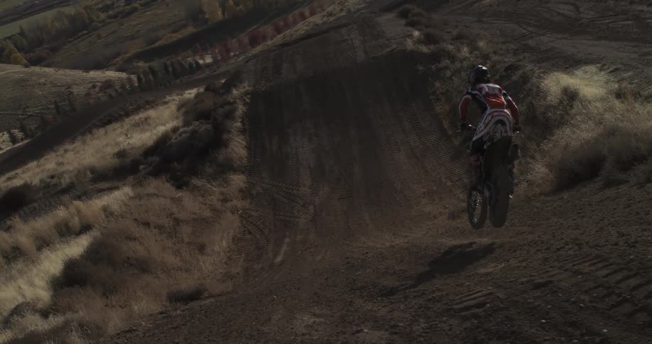 Super slow motion from behind a motocross rider as they fly through the air over a large dirt jump and land perfectly