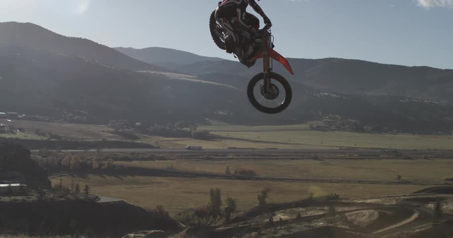 A motocross rider launches into the air in epic slow motion, suspended against the blue sky with a mountain valley below
