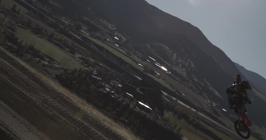 A motocross rider performs a spectacular whip trick in super slow motion over a dirt track with a scenic mountain valley in the background