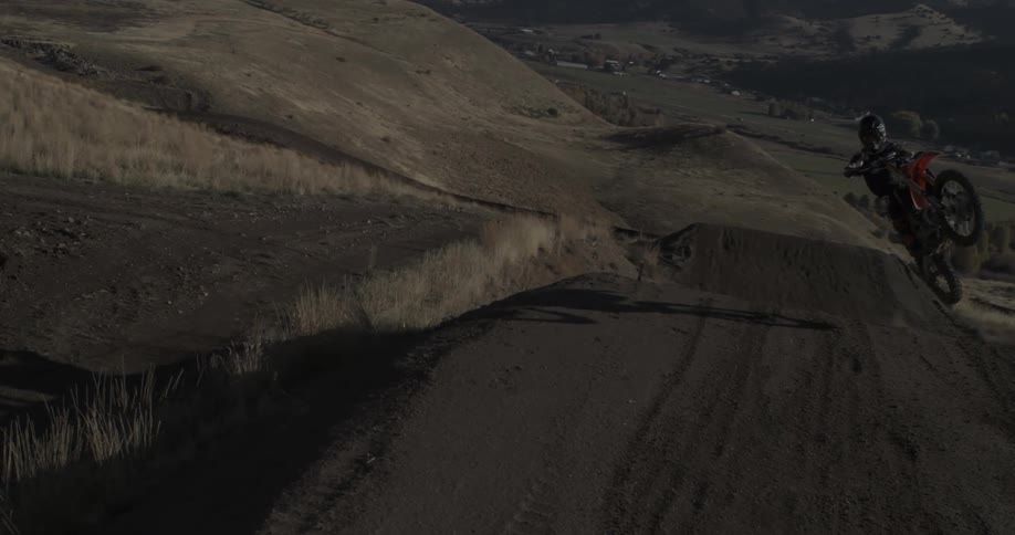 A motocross rider performs a high-flying jump off a dirt ramp in a hilly, rural landscape, captured in dramatic slow motion