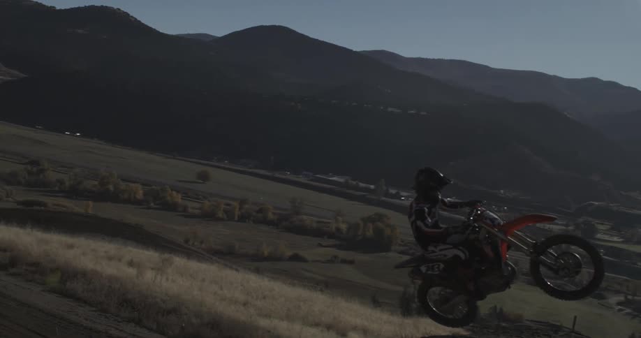 In stunning slow motion, a motocross rider performs a wheelie off a dirt jump, soaring through the air with majestic mountains in the background