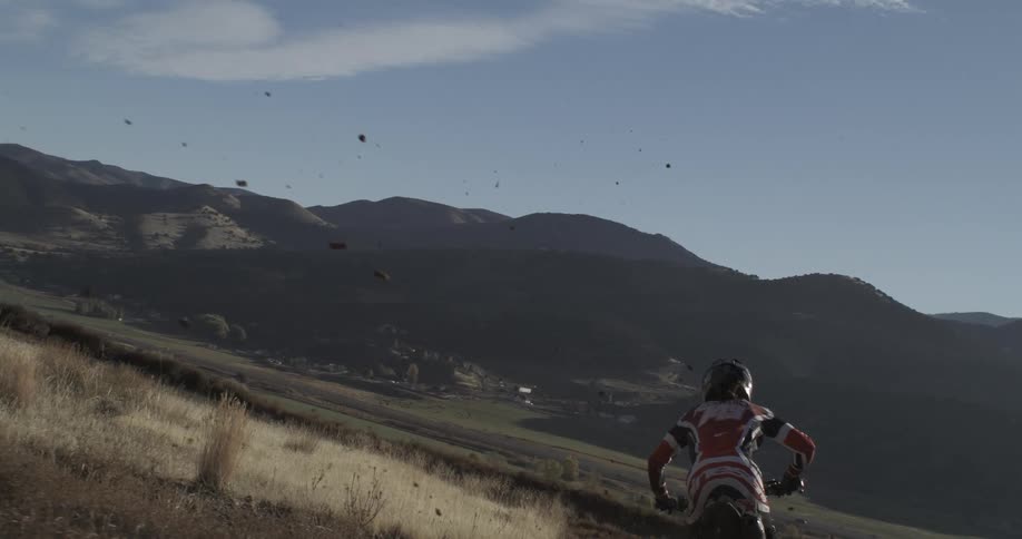 Dirt flies up in slow motion from behind a motocross rider poised on a mountain trail overlooking a scenic valley
