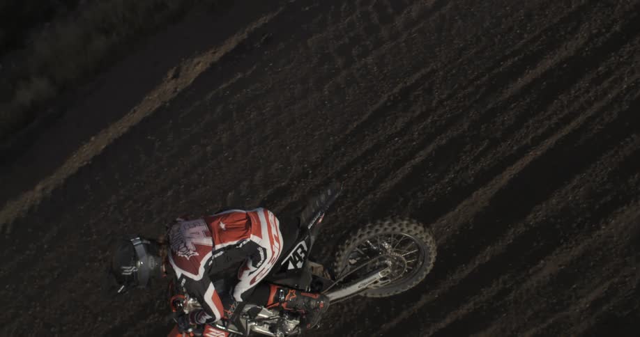 An overhead slow-motion shot captures a motocross rider in full gear accelerating on a dirt track, kicking up dust as they speed away