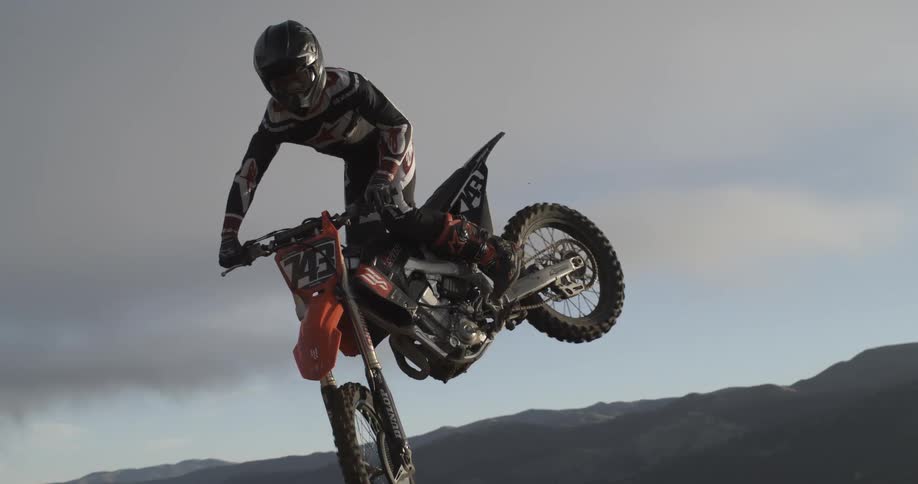A professional motocross rider performs a jump, soaring through the air in dramatic super slow motion against a backdrop of rolling hills
