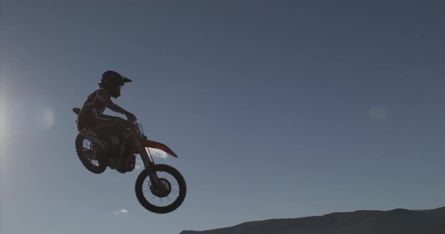 A motocross rider is captured in extreme slow motion as they fly through the air, silhouetted against the sun over a vast mountain landscape