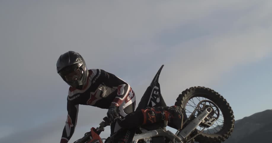 An extreme motocross rider performs a stylish jump in super slow motion, captured from a low angle against the sky and a mountain landscape