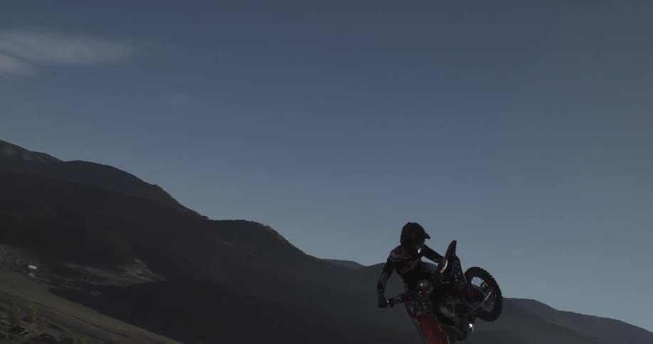 A motocross rider jumps in slow motion with a mountain range and bright sun in the background