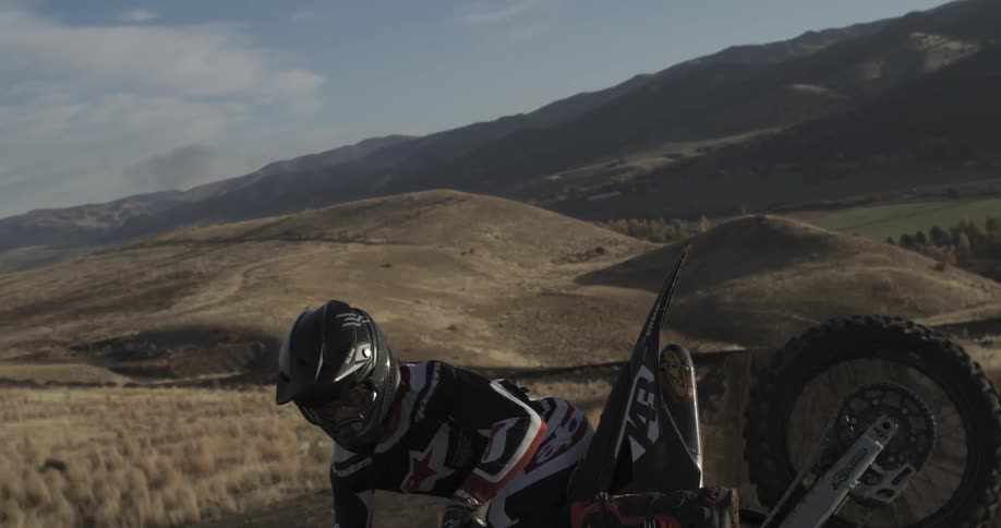 A motocross rider hangs in the air mid-jump in super slow motion, set against a backdrop of rolling hills
