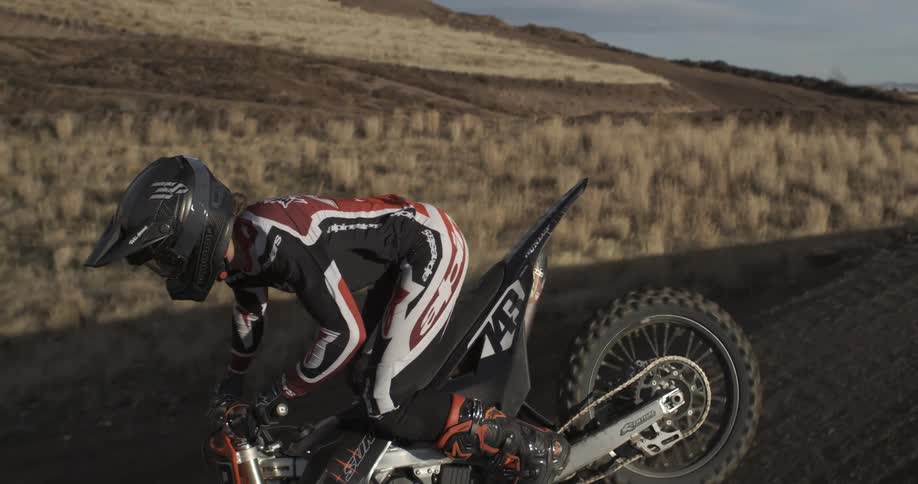 Slow motion pan reveals a motocross rider in full gear sitting on their dirt bike on a rugged, hilly course