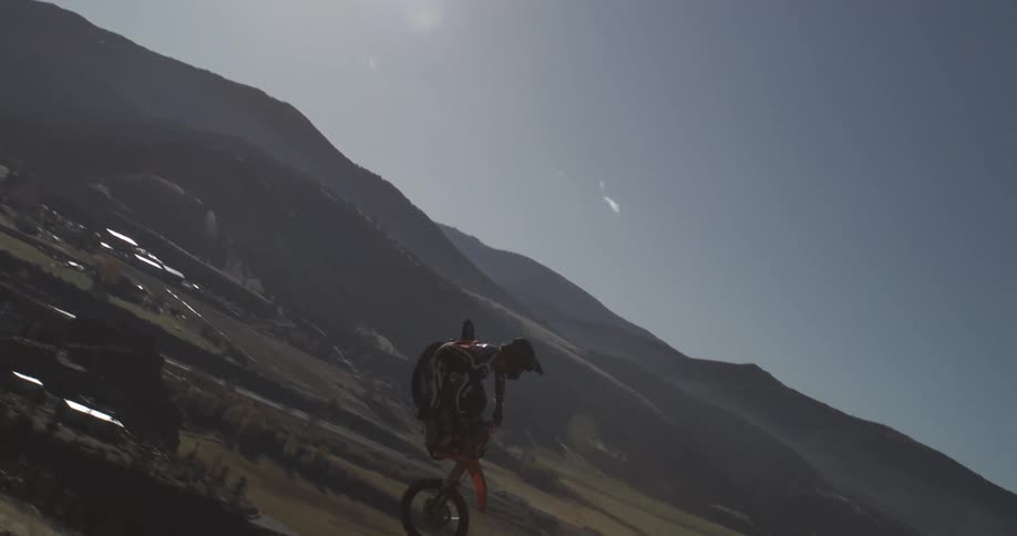 An extreme motocross rider performs a spectacular mid-air stunt in super slow motion, silhouetted by the sun over a mountain landscape