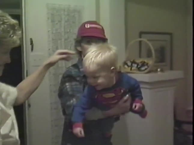 Vintage VHS footage shows a happy baby in a Superman costume laughing while being held by his mother