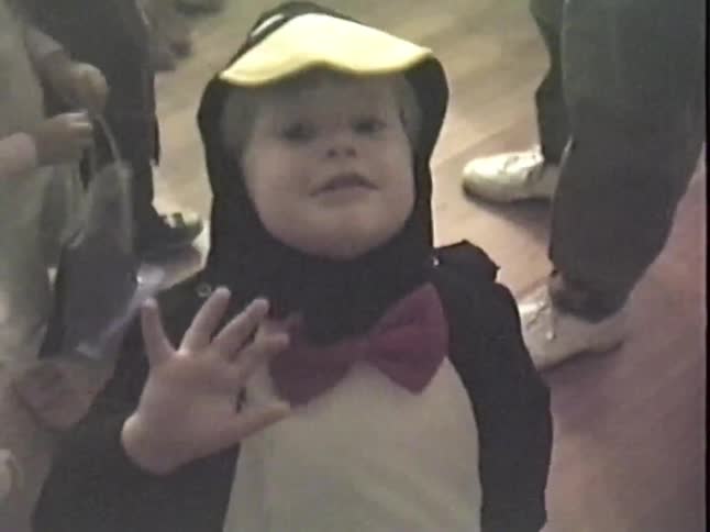 A cute toddler in a penguin Halloween costume waves at the camera before dropping their candy pail