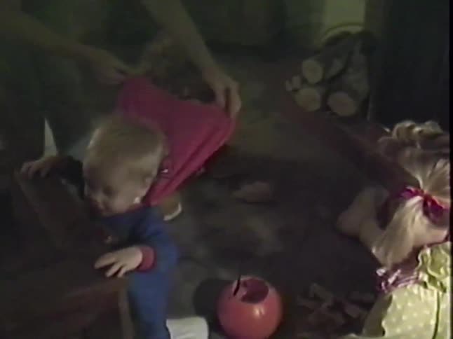 A toddler in a Superman costume smiles as an adult helps another child with their Halloween costume
