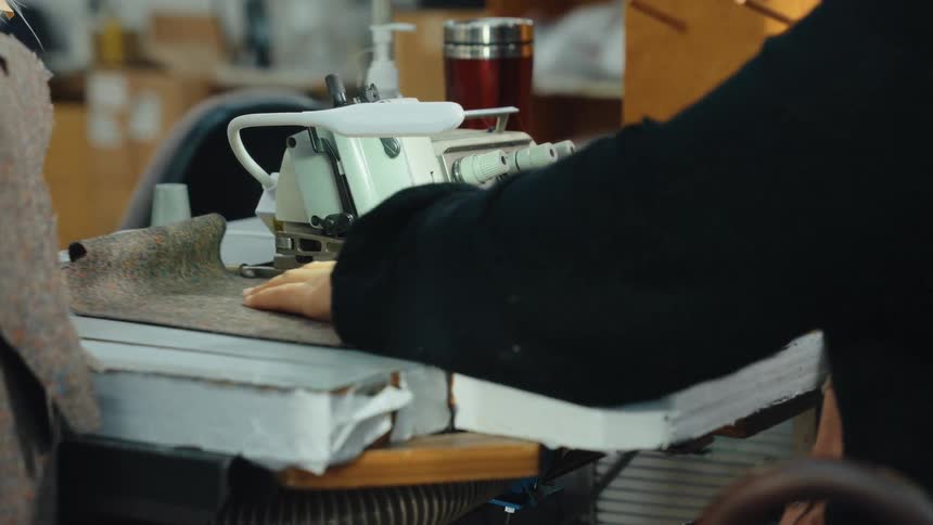 A seamstress skillfully operates an industrial overlock sewing machine to stitch the edge of a piece of thick fabric in a workshop