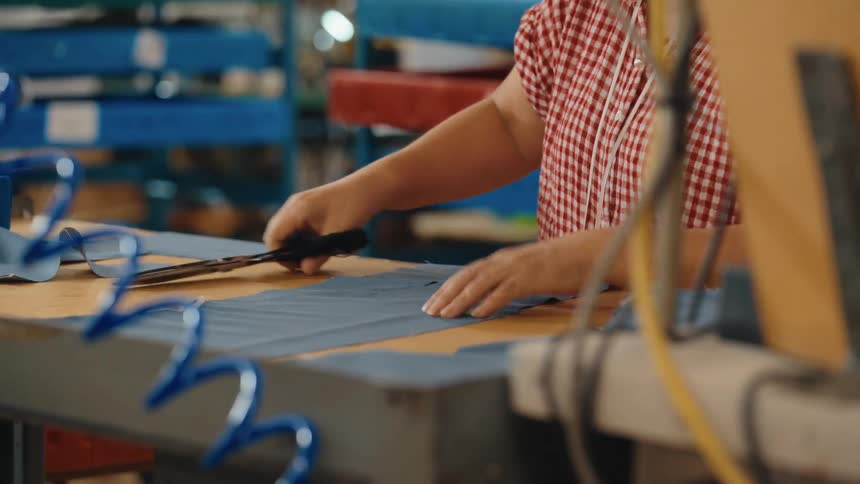 A seamstress carefully cuts a piece of blue fabric with large scissors at a workbench in a factory