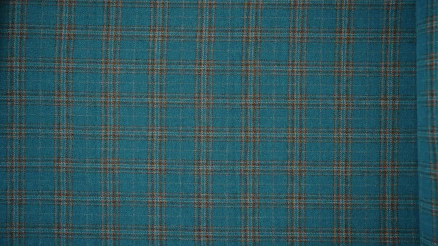 A hand unrolls a bolt of teal and brown plaid fabric, revealing its full pattern from a top-down perspective