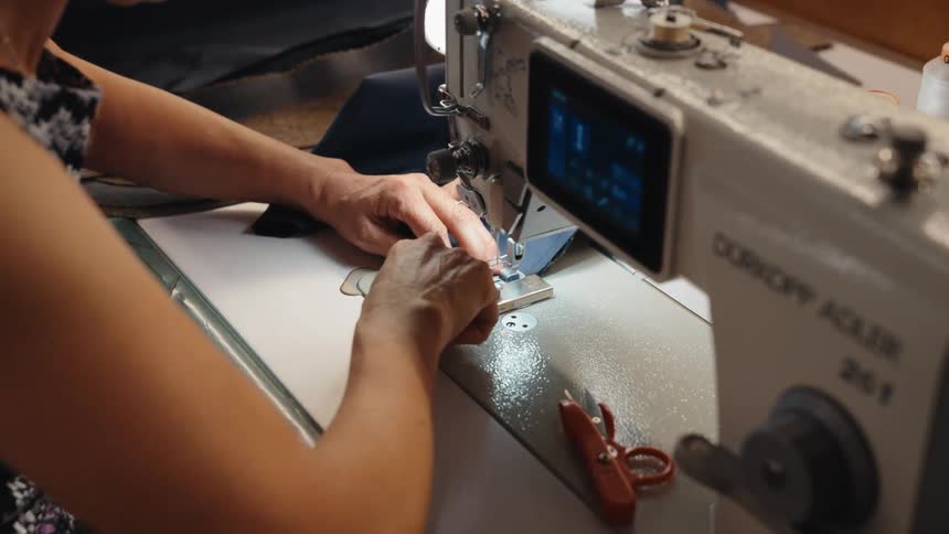 Over the shoulder shot of a seamstress carefully guiding blue fabric through an industrial sewing machine