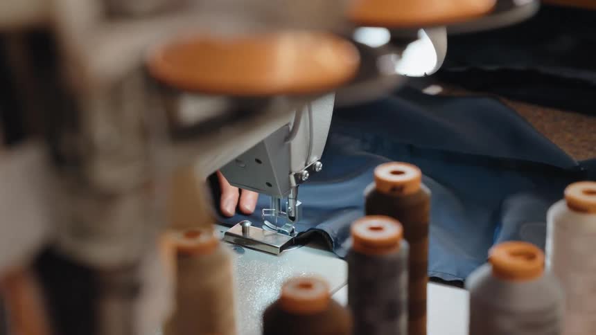 Close-up of a sewing machine stitching blue fabric in a textile workshop with spools of thread in the foreground