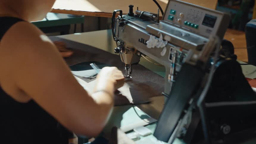 An over-the-shoulder shot of a seamstress operating an industrial sewing machine in a sunlit workshop