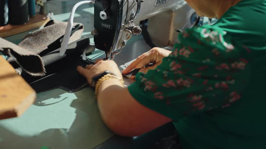Over-the-shoulder view of a seamstress carefully guiding dark fabric through an industrial sewing machine in a sunlit workshop