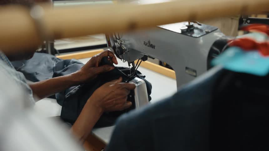 A garment worker's hands skillfully guide dark fabric through an industrial sewing machine in a workshop