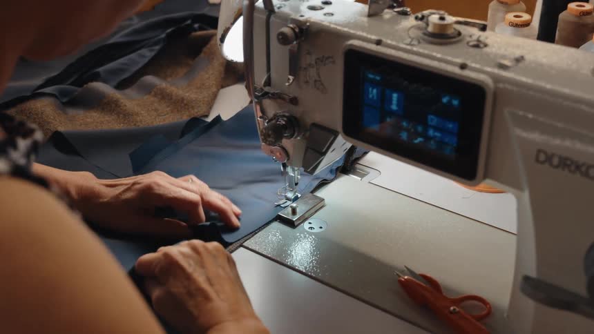An over-the-shoulder view shows a skilled seamstress using a modern industrial sewing machine to stitch a piece of dark blue fabric