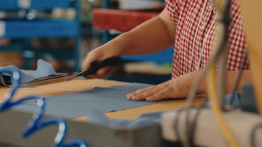 A factory worker wearing a checkered shirt meticulously cuts a piece of blue fabric with large scissors in slow motion